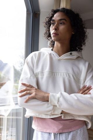 Non-binary person standing by window, arms crossed, gazing thoughtfully in natural light