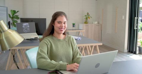 Smiling woman working on laptop in bright modern home office with plants and floorplan