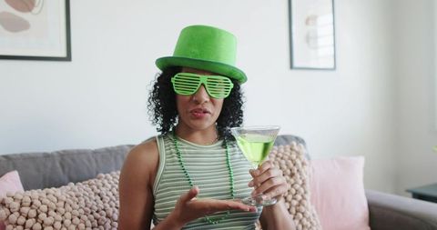African woman celebrating with green cocktail, wearing festive hat and neon shutter shades