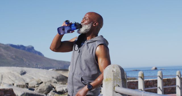 Senior man drinking water after intense workout by seaside. Ocean and mountains in the background highlight natural beauty. Perfect for ads about healthy living, hydration, fitness motivation, and senior wellness.