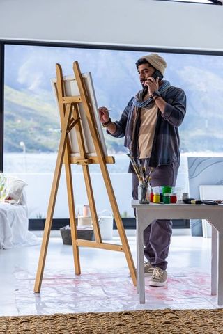 Painter talking on phone while painting at easel in waterfront studio with mountain view