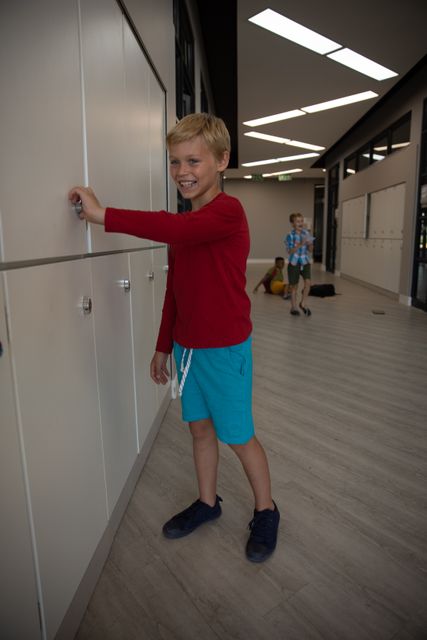 Smiling caucasian elementary schoolboy opening locker while standing in corridor. unaltered, education, childhood and school concept.
