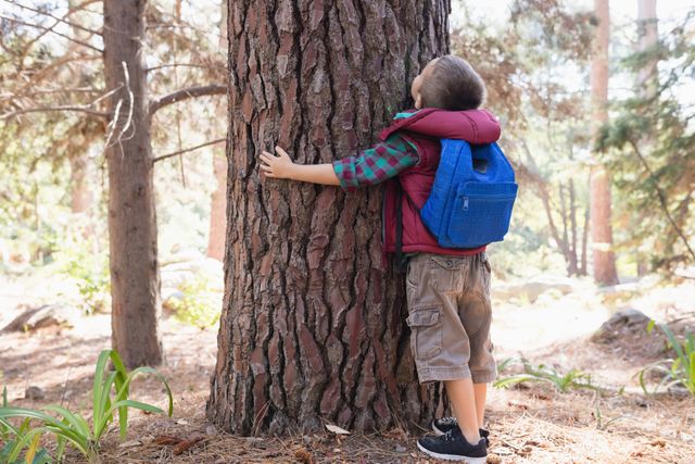 Rear view of boy with backpack embracing tree in forest