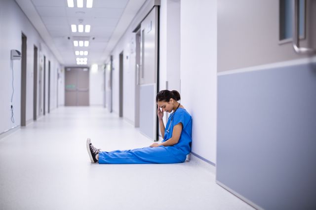 Female doctor in blue scrubs sitting on the floor of a hospital corridor, appearing stressed and exhausted. Ideal for illustrating themes of healthcare worker burnout, mental health challenges in the medical field, and the pressures faced by medical professionals. Useful for articles, blogs, and campaigns focusing on healthcare, mental health awareness, and workplace stress.