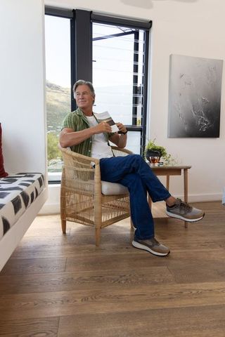 Middle-aged man reading magazine while relaxing in rattan chair beside louvered window