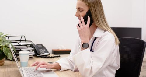 Female physician talking on phone while typing patient notes on laptop clinic office
