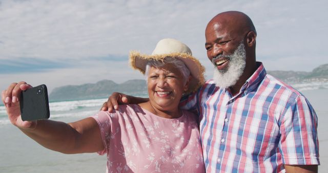 A cheerful senior couple enjoying time at the beach, capturing memories by taking a selfie. Ideal for use in travel promotions, retirement lifestyle articles, and advertisements focusing on vacation for seniors.