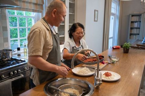 Asian senior couple preparing noodles with tomato sauce on wooden kitchen island