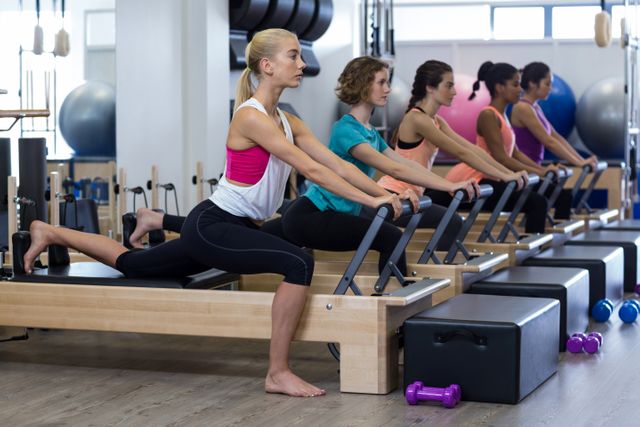Group of women engaging in a Pilates workout using reformer machines in a modern gym. Ideal for illustrating fitness classes, active lifestyles, and group training sessions. Useful for marketing fitness programs, showcasing gym equipment, and promoting health and wellness activities.