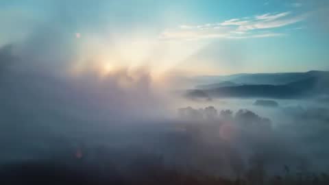 Drone Elevation Through Misty Forest Valley at Sunrise Showing Sunbeams and Lens Flare