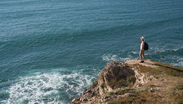 Explorer standing on rocky cliff gazing over vast ocean with backpack and brimmed hat