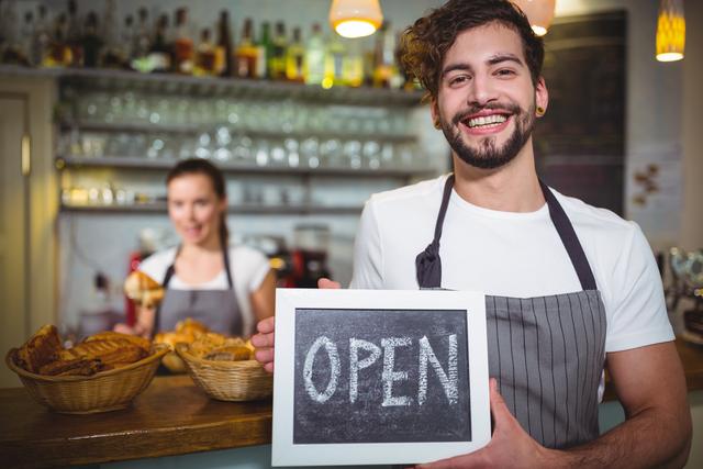 This image shows a cheerful waiter holding an open sign in a cozy cafe. The background features another staff member and a display of pastries, creating a welcoming atmosphere. Ideal for use in marketing materials for cafes, restaurants, and small businesses to convey friendliness and excellent customer service.