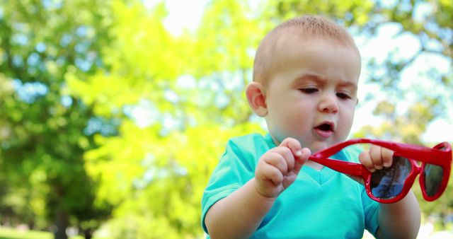 Baby playing with red sunglasses in a green park. Perfect for themes related to childhood, curiosity, outdoor activities, and summer fun. Suitable for use in parenting blogs, child care promotions, and advertisements focusing on family outings.