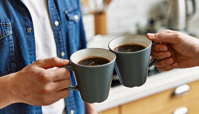 Two adults clinking gray ceramic mugs over kitchen counter morning coffee toast, cozy