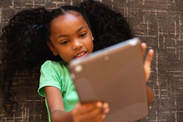 Young African American girl lying on carpet using digital tablet. Ideal for educational content, technology use in children, online learning resources, and childhood relaxation themes. Suitable for articles on digital literacy, child development, and home learning environments.