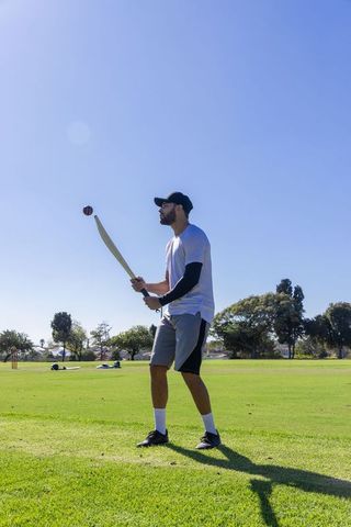 Man practicing cricket batting on sunny park field with ball midair
