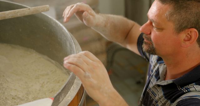 Middle-aged baker mixing flour manually in large container in bakery kitchen. Flour visible on hands and container, which indicates intensive preparation process. Useful for showcasing traditional baking methods, artisan bread making, and recipes or articles focused on bakeries and culinary arts.