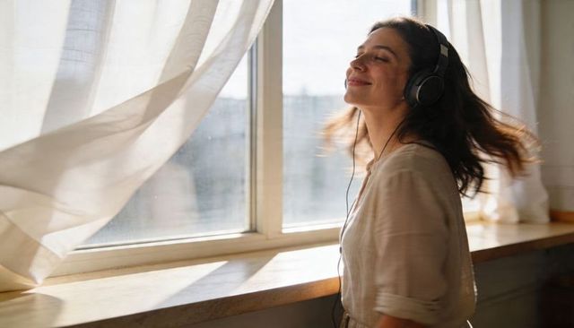 Woman listening to music wearing headphones by sunlit window with flowing curtains smiling