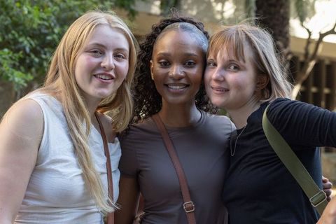 Smiling diverse female friends posing and leaning together on sunlit campus courtyard