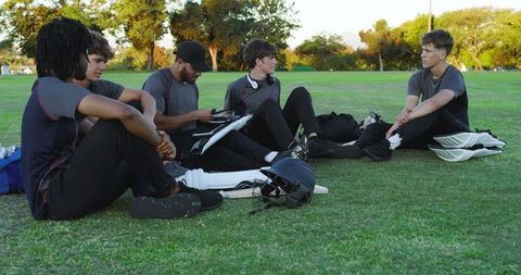 Youth cricket teammates sitting on grass reviewing strategy on tablet with helmets and pads outdoors