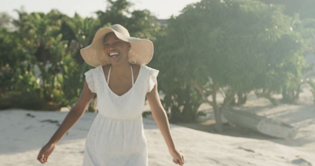 Young woman wearing white dress and sun hat, walking and laughing on a sunny beach. Trees and plants in background, suggesting tropical location. Perfect for travel advertisements, summer promotions, lifestyle blogs, or wellness content.