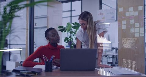 Two Women Collaborating on Strategy at Modern Office Desk with Glass Partition, Charts