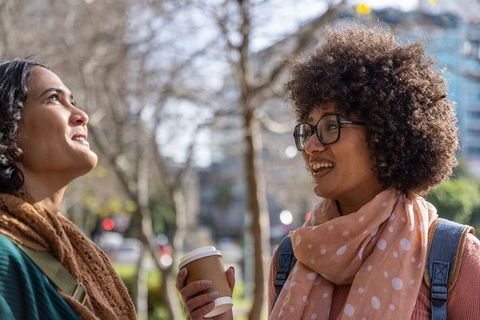 Two friends chatting and laughing on sunny urban park walkway holding takeaway coffee