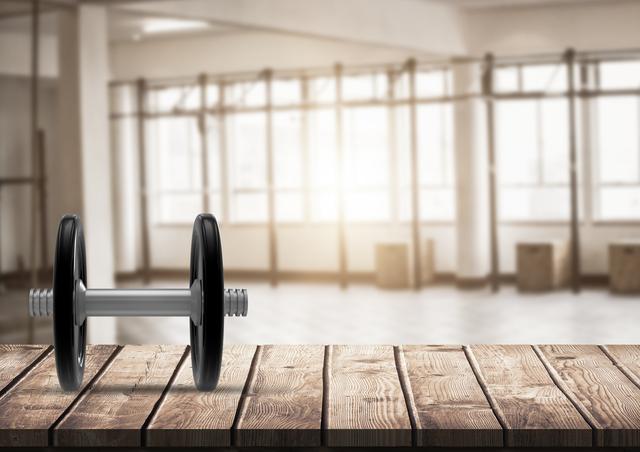 Dumbbell resting on wooden plank in gym with bright, sunlit background. Useful for fitness blogs, workout posters, or health-related materials promoting strength training and gym interior aesthetics.