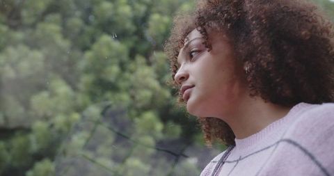Contemplative woman gazing through rain-speckled glass, purple sweater, blurred greenery
