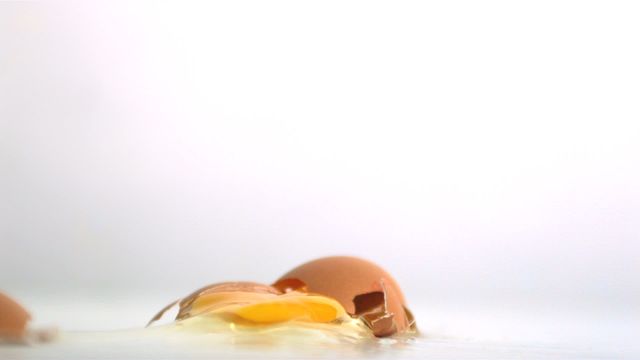 Close-up view of a broken egg with scattered shell pieces and exposed yolk on a white surface. Useful for illustrating concepts of food, cooking, or kitchen accidents. Suitable for food blogs, cooking tutorials, educational materials, or marketing content related to kitchen products.