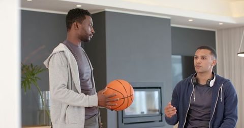 Diverse young men holding basketball and talking in modern living room, casual friendship