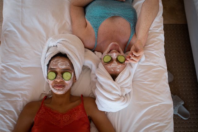 Two diverse female friends are lying on a bed with face masks and towels wrapped around their heads. They have cucumber slices on their eyes, enjoying a relaxing spa day together. This image can be used for promoting skincare products, wellness retreats, beauty treatments, or articles about self-care and friendship.