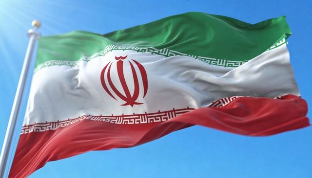 Iranian flag waving in wind against blue sky with sun flare, kufic calligraphy and emblem