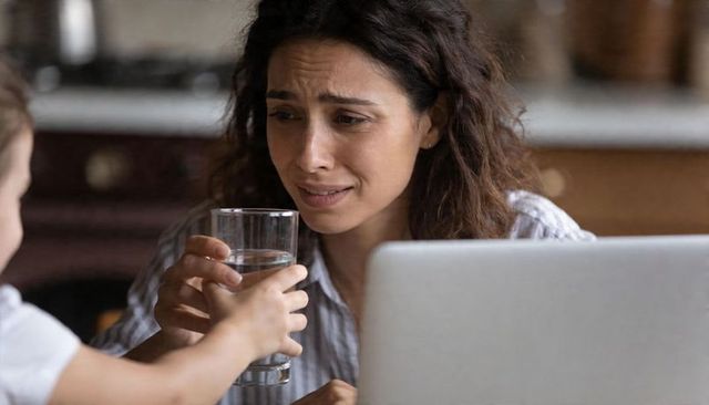 Tired mother accepting glass of water from child while working remotely at kitchen table