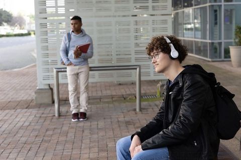 Teen students listening to music on campus bench while classmate holding notebook