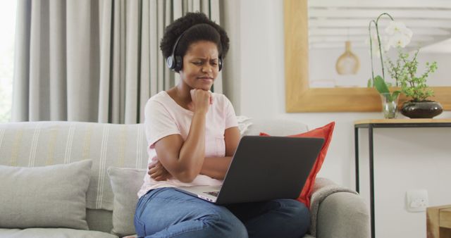 African American woman wearing headphones and using a laptop while sitting on a couch at home. She appears focused and engrossed, which makes it suitable for uses related to remote work, e-learning, or casual internet browsing. It can also be used in articles or advertisements about using technology for work from home, relaxation with media, or modern living.