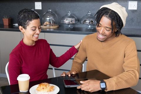 African american couple sharing tablet and croissant while smiling at cozy cafe breakfast