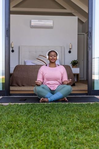 African american woman meditating at sliding door listening to headphones outdoors