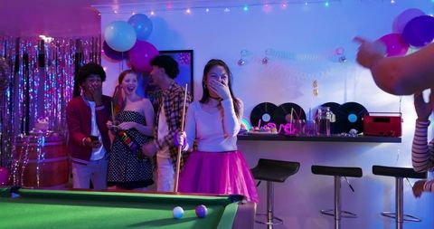 Friends celebrating at neon game room playing pool with asian woman in pink tulle skirt