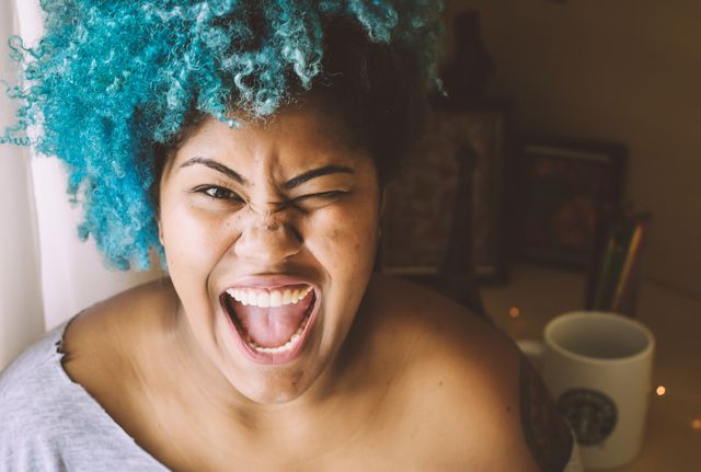 Woman with vibrant blue curly hair winking and smiling joyfully. Woman exuding happiness and energy. Can be used in lifestyle blogs, advertisements, and social media posts to convey fun, individuality, and positive emotions.