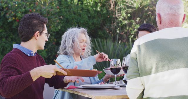 Family with multi-generational members enjoying meal outside in sunny backyard. Includes older adults and younger adults gathered around table sharing food and conversation. Ideal for concepts of family bonding, outdoor dining, social gatherings, and summer activities.