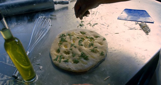 Chef's hand sprinkling fresh herbs on focaccia dough on work surface with whisk and olive oil bottle. Great for use in cooking tutorials, baking blogs, and culinary magazine features. Perfect for showcasing artisanal and homemade food preparation.