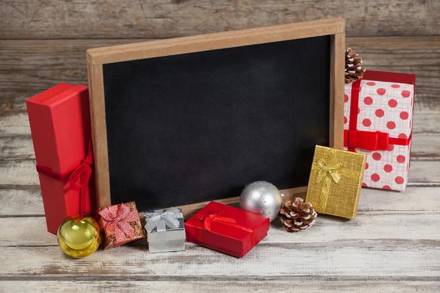 Arrangement depicts wrapped Christmas gifts and festive decorations around a blank chalkboard on wooden surface. Chalkboard can be used for messages or holiday greetings. Ideal for Christmas promotions, holiday invitations, advertisements, and seasonal greeting cards.