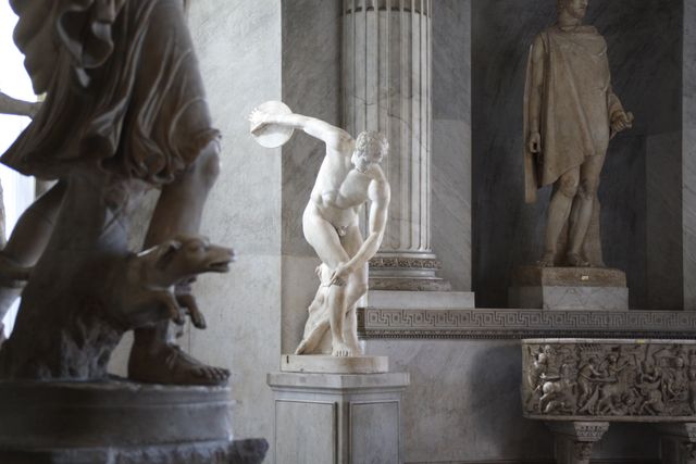 A collection of ancient sculptures made of marble displayed in a historical museum. The statues depict classical figures, including a man holding a disc and other mythological scenes carved in bas-relief. Ideal for use in historical art presentations, cultural heritage documentaries, and educational materials about classical art and history.