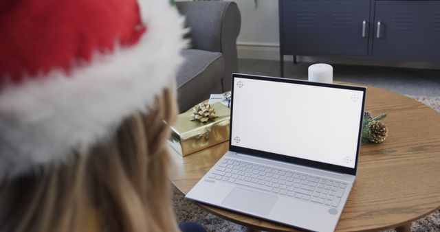 Woman in Santa hat having a video call on laptop during Christmas season. Gift and candle on coffee table, creating festive home atmosphere. Ideal for use in articles or ads about holiday season, virtual celebrations, or remote communications.