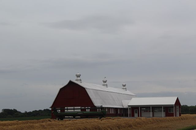 Traditional red barn with white roof and silos stands prominently in a field recently harvested. The overcast sky adds a moody atmosphere to the rural landscape. Perfect for depicting American farm life, agricultural themes, or rustic countryside living.