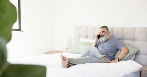 Senior Man Relaxing on Bed with Laptop and Smartphone in Modern Bright Bedroom