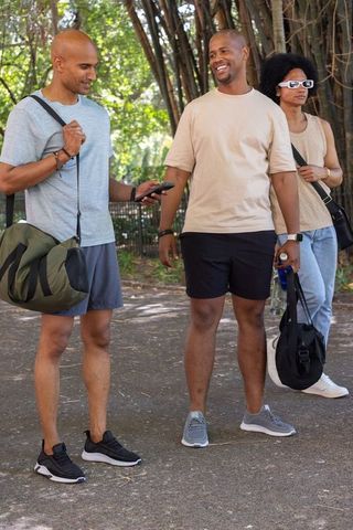 African american friends sharing phone after workout, holding gym bags on tree-lined park path