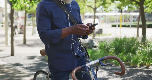 Middle-aged man standing outdoors with a bicycle, using his smartphone while listening to music through earphones. Ideal for themes related to urban lifestyle, communication, casual fashion, and outdoor activities. Great for illustrating modern technology use and cycling in the city within green surroundings.