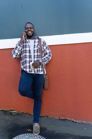 African american man leaning on urban wall, talking on phone, holding coffee, casual look